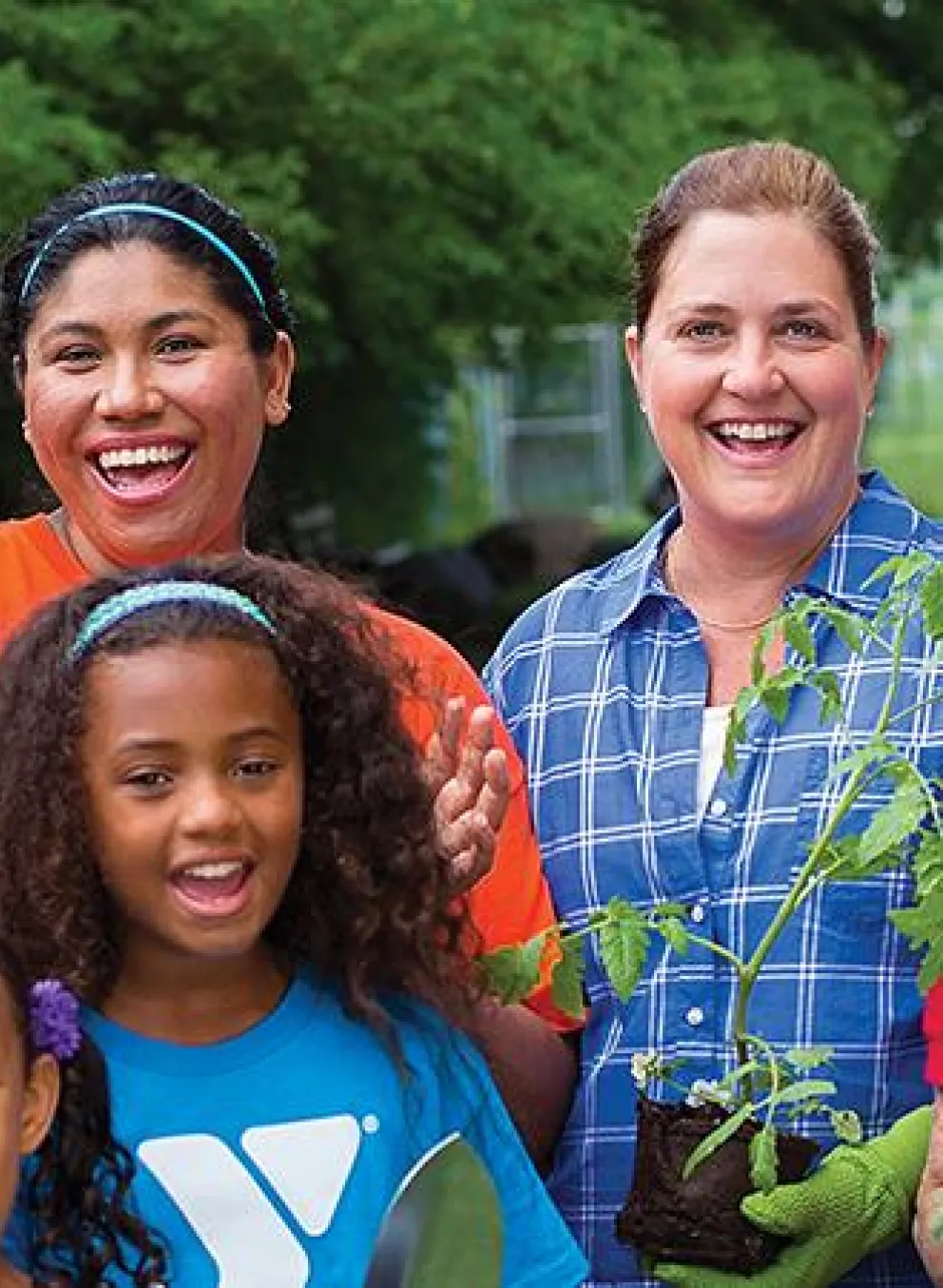 A diverse group of smiling adults and children stand together, holding young tomato plants, possibly involved in a community gardening project.