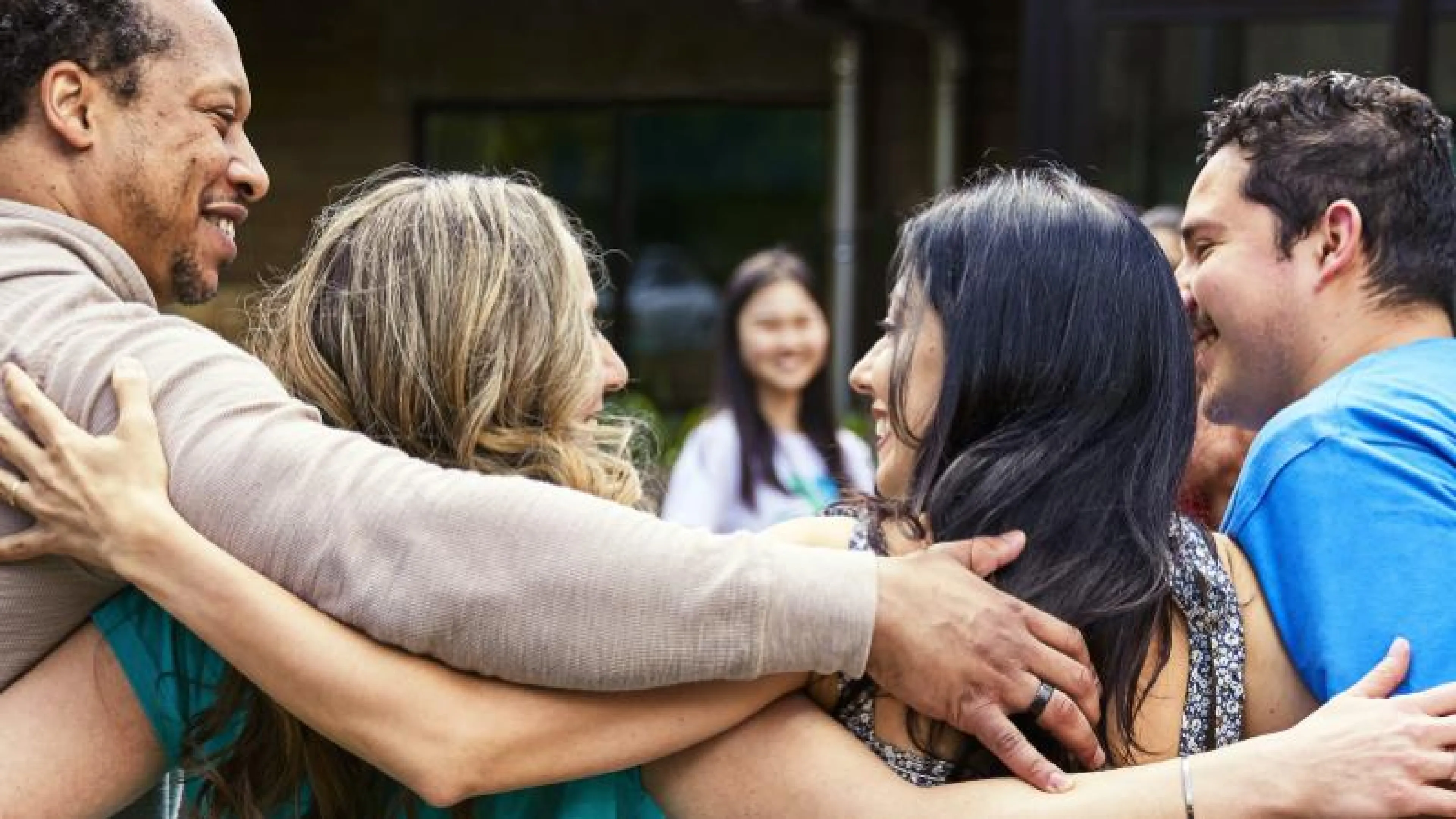 Four smiling adults in casual clothes embrace, arms around shoulders, standing outside a building.