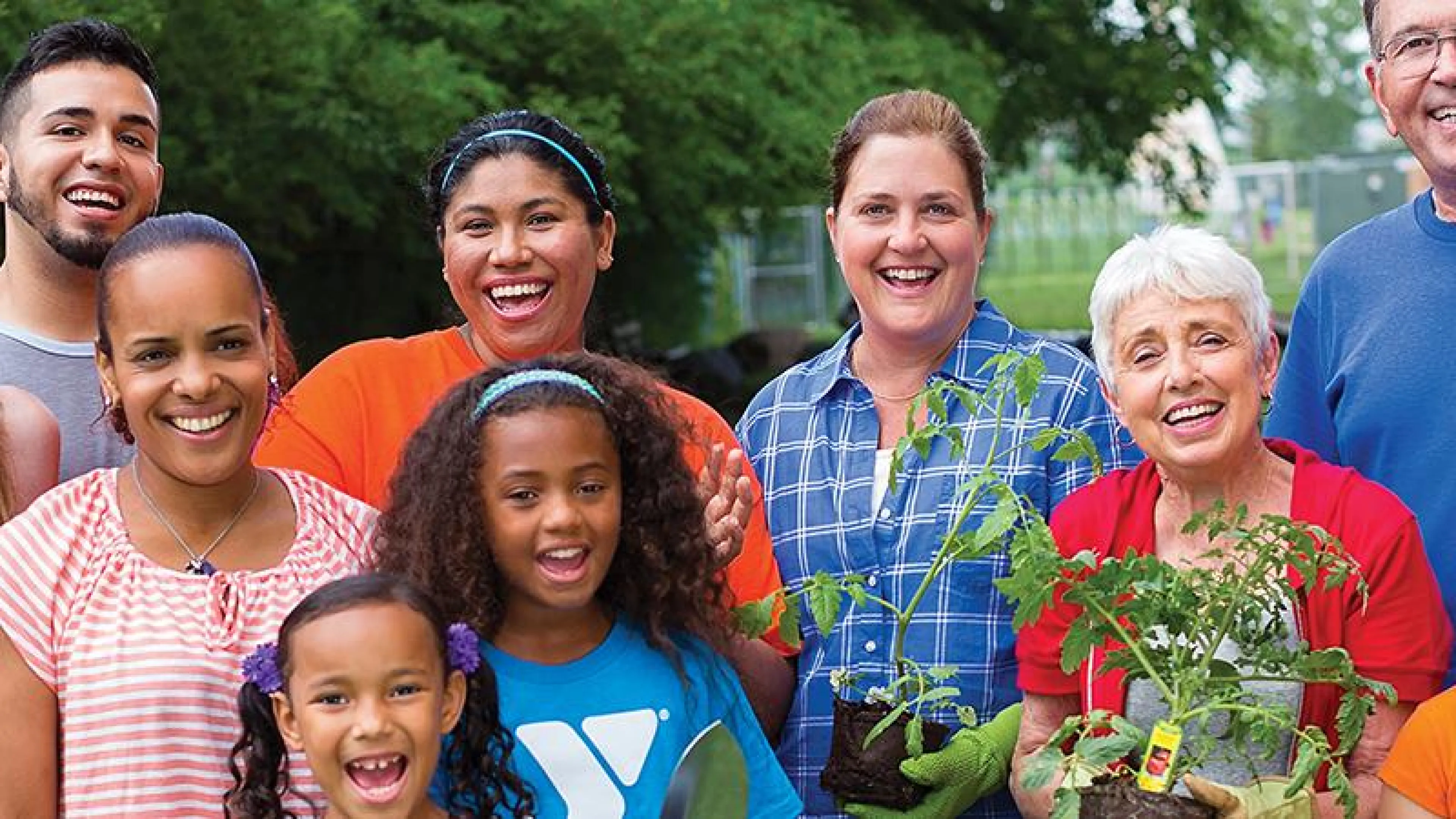 A diverse group of smiling adults and children stand together, holding young tomato plants, possibly involved in a community gardening project.